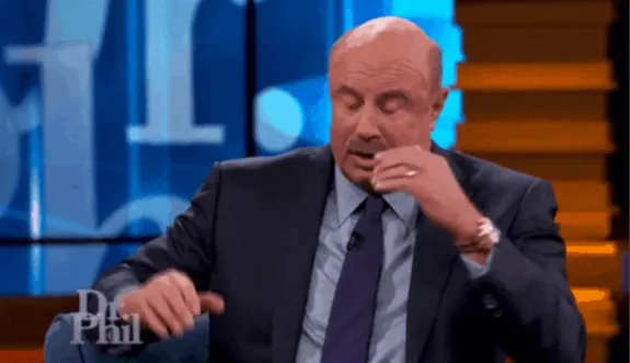 Bald man in a suit sitting on a talk show set gesturing while talking.