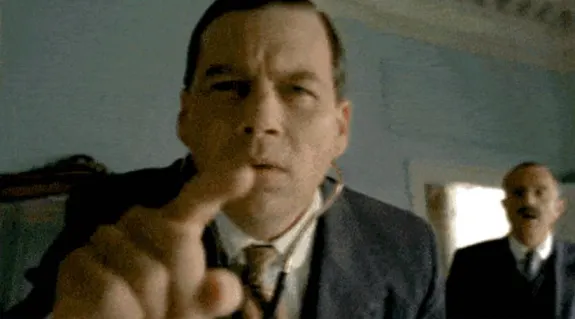 Man in a suit pointing at the camera indoors with another person in the background.