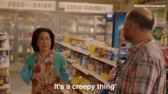 Woman wearing apron and gloves confronting man in grocery store aisle.