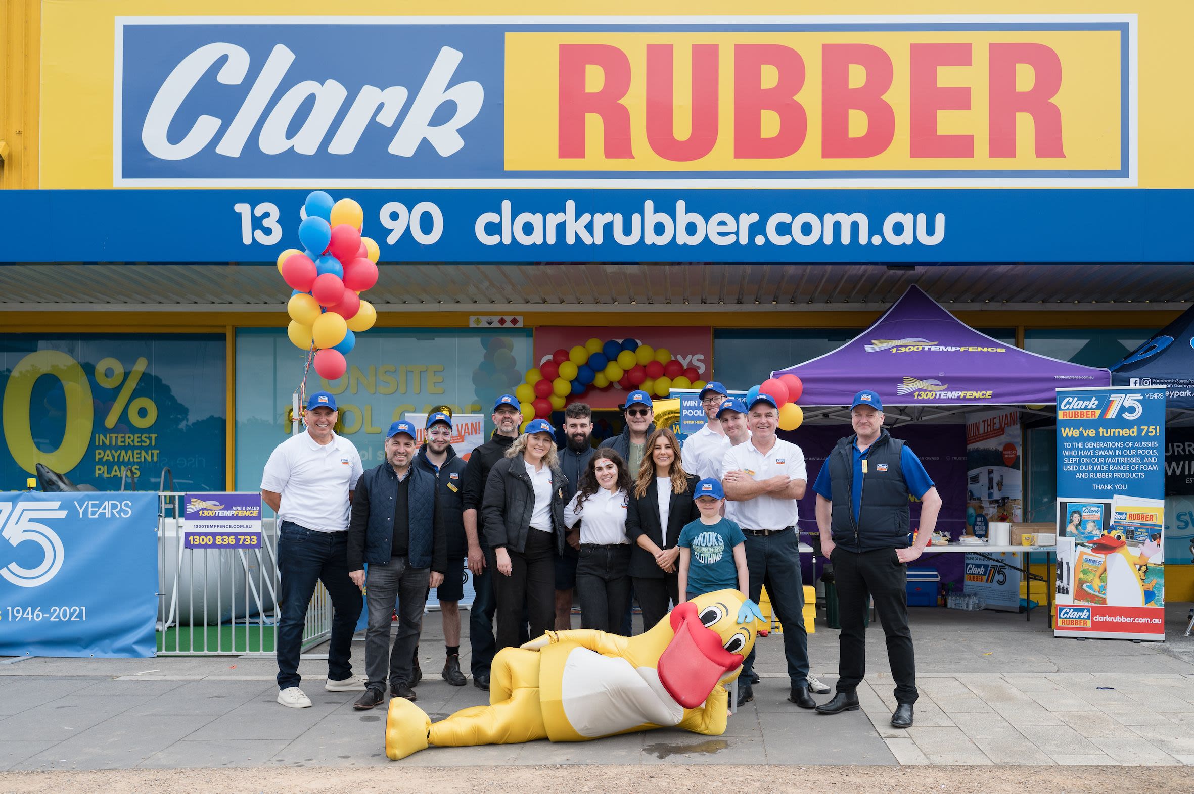 Group of people celebrating Clark Rubber's anniversary at store entrance with balloons and mascot.