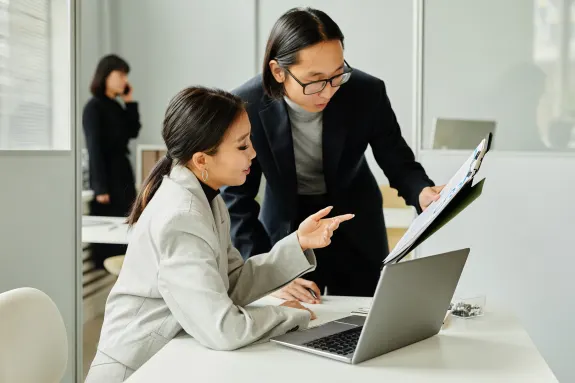 Two business professionals reviewing documents at a desk with a laptop in an office setting.