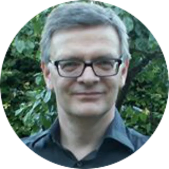 Man wearing glasses and a dark shirt smiling outdoors with greenery in background.