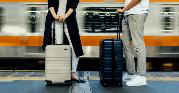 Two travelers with suitcases waiting at a train station with a passing train.