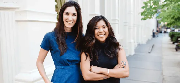 Two women smiling and standing outdoors with an urban background.
