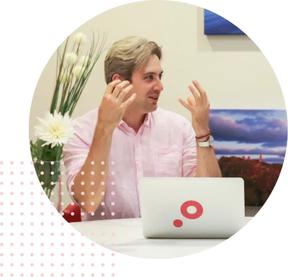 Person in pink shirt talking animatedly at a desk with a laptop